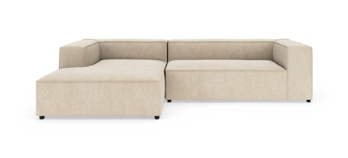Beige modulsofa i L-form med bred chaiselong, lav profil og kantede armlæn, frilagt på hvid baggrund.