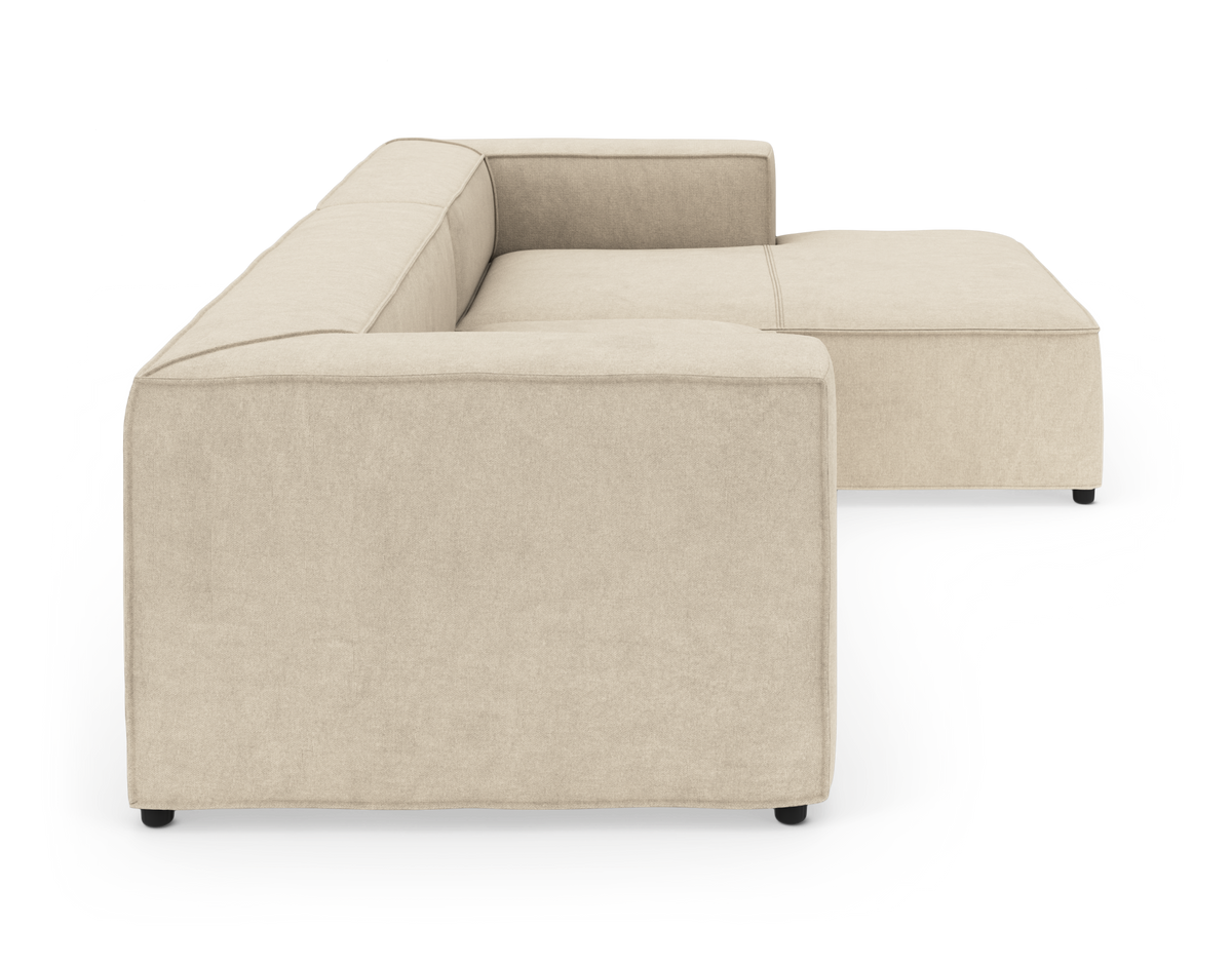 Beige modulsofa i L-form med brede armlæn, set fra siden på hvid baggrund.