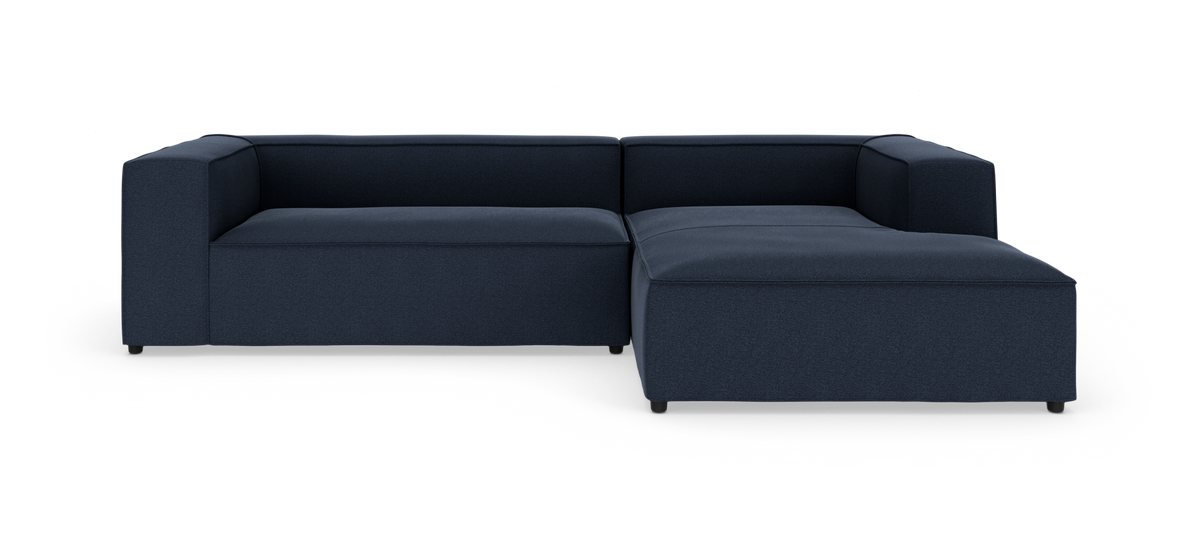 Mørkeblå modulær L-formet sofa med chaiselong til højre, lave blok-armlæn og stofbetræk, frilagt på hvid baggrund.