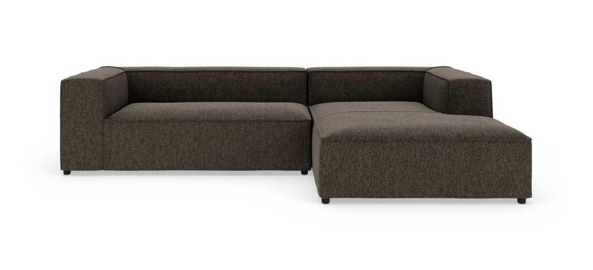 Mørkegrå modulær L‑sofa på hvid baggrund med lav ryg og bred chaiselong til højre.