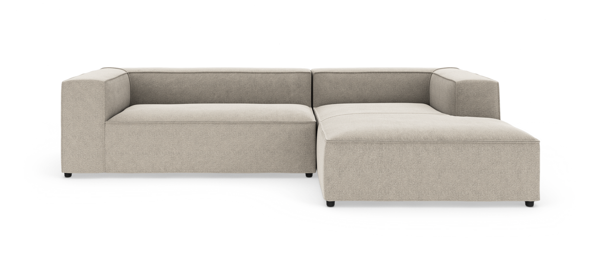 Modulær L-formet hjørnesofa i lys beige stof med brede blokarmlæn og højrevendt chaiselong, frilagt på hvid baggrund.