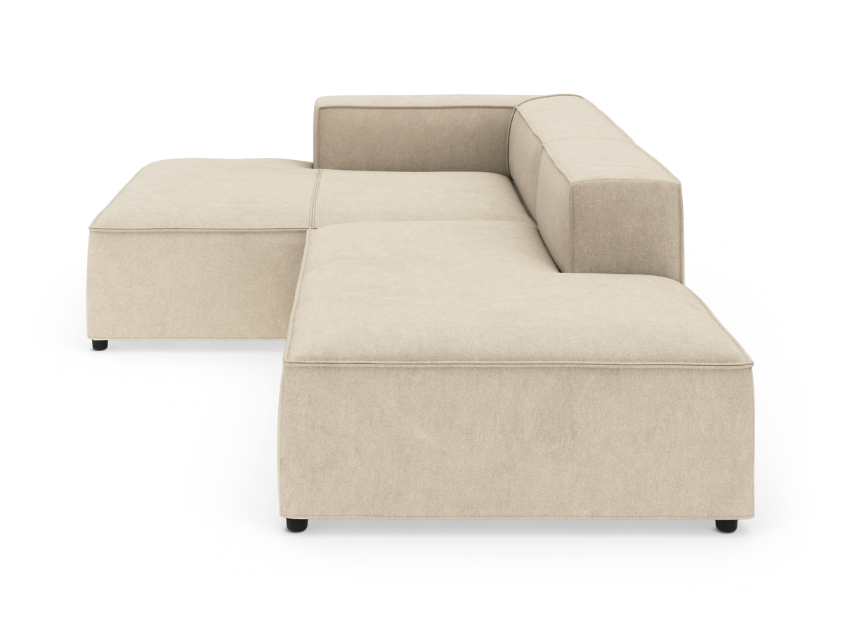 Beige modulsofa i L‑form med brede sæder og lave sorte ben, frilagt på hvid baggrund.