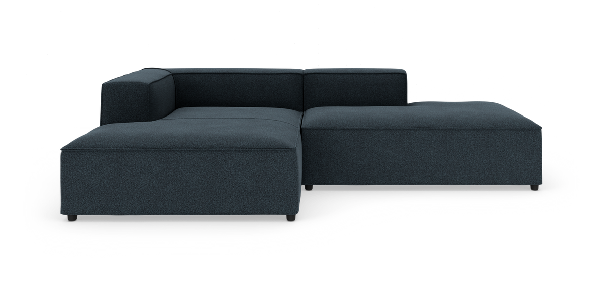 Mørkeblå modulsofa i L‑form med lav ryg og brede sædehynder, fotograferet forfra i studio.