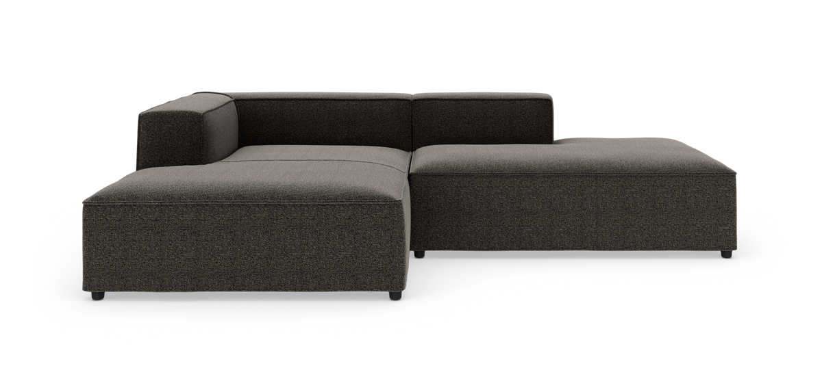 Modulær L‑formet sofa i mørkegråt stof med lav ryg og venstre armlæn, frilagt på hvid baggrund.