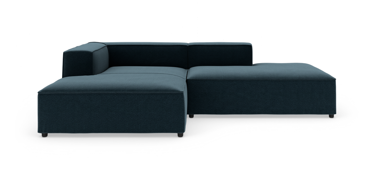 Modulær hjørnesofa i mørk blågrøn tekstil med lav ryg og chaiselong, frilagt på hvid baggrund.