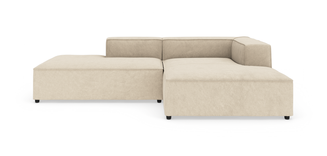Beige modulsofa i L‑form med brede armlæn og lav profil på hvid baggrund.