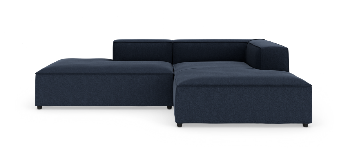 Mørkeblå, modulær L‑formet sofa med lav profil og bred chaiselong til venstre, kubeformede armlæn; fristående produktbillede på hvid baggrund.