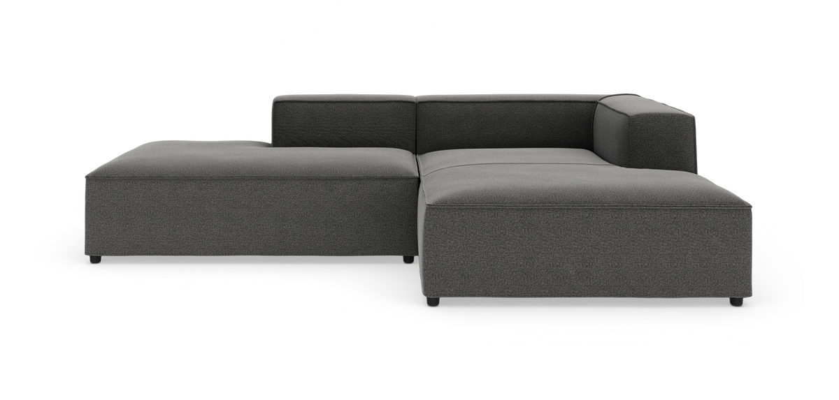 Mørkegrå modulsofa i L‑form med chaiselong og lave blok-armlæn, fotograferet i studie mod hvid baggrund.
