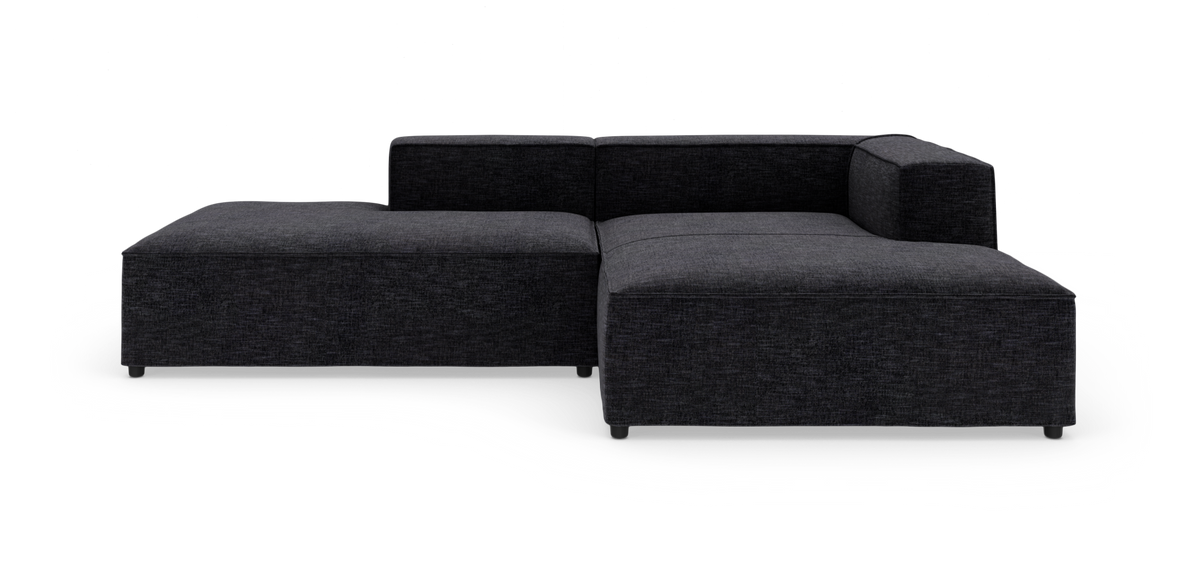 Mørkegrå modulsofa i L‑form med lav ryg og brede armlæn, fotograferet mod hvid studiobaggrund.