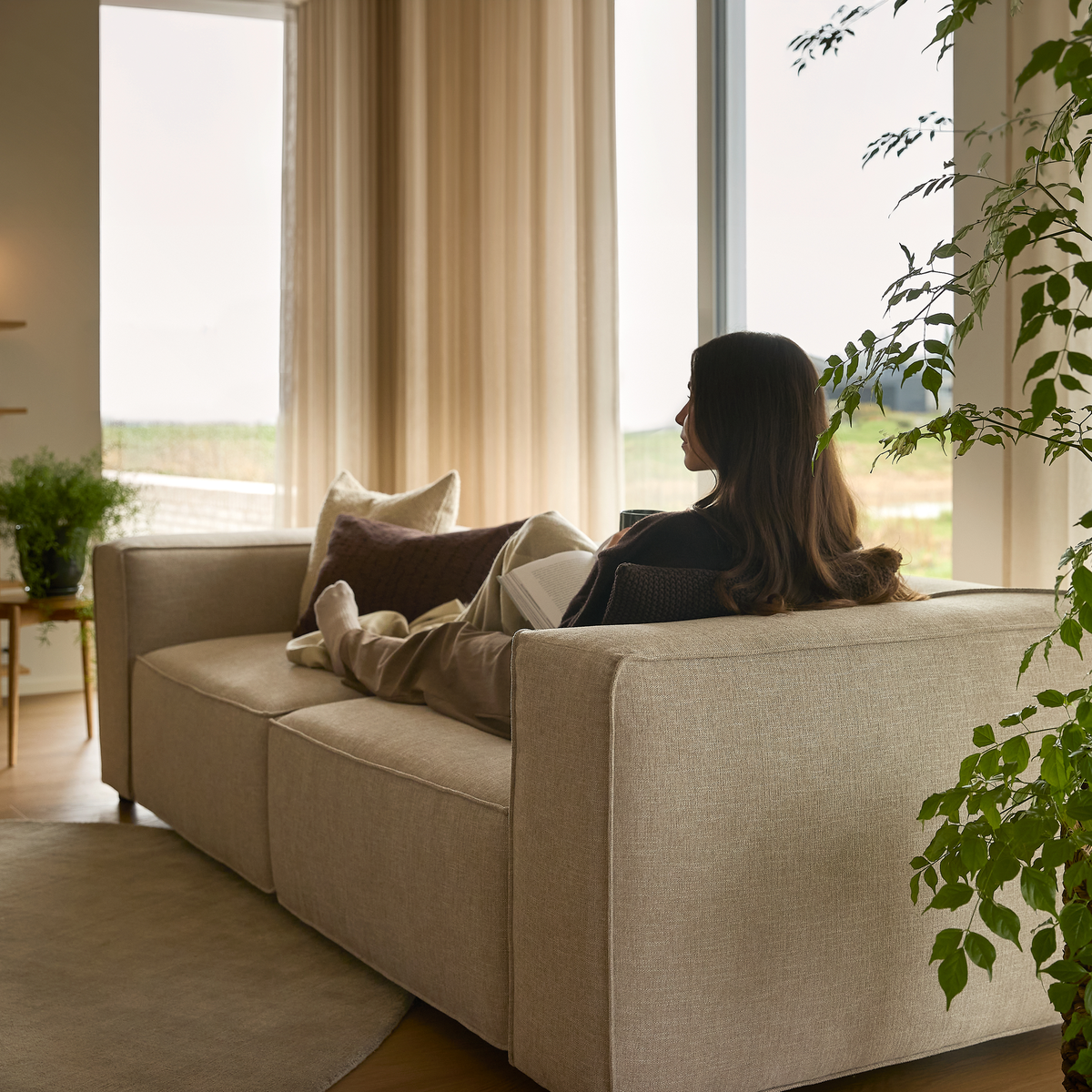Person ligger og læser på beige sofa ved stort vindue, neutrale toner, grønne planter og lyse gardiner.