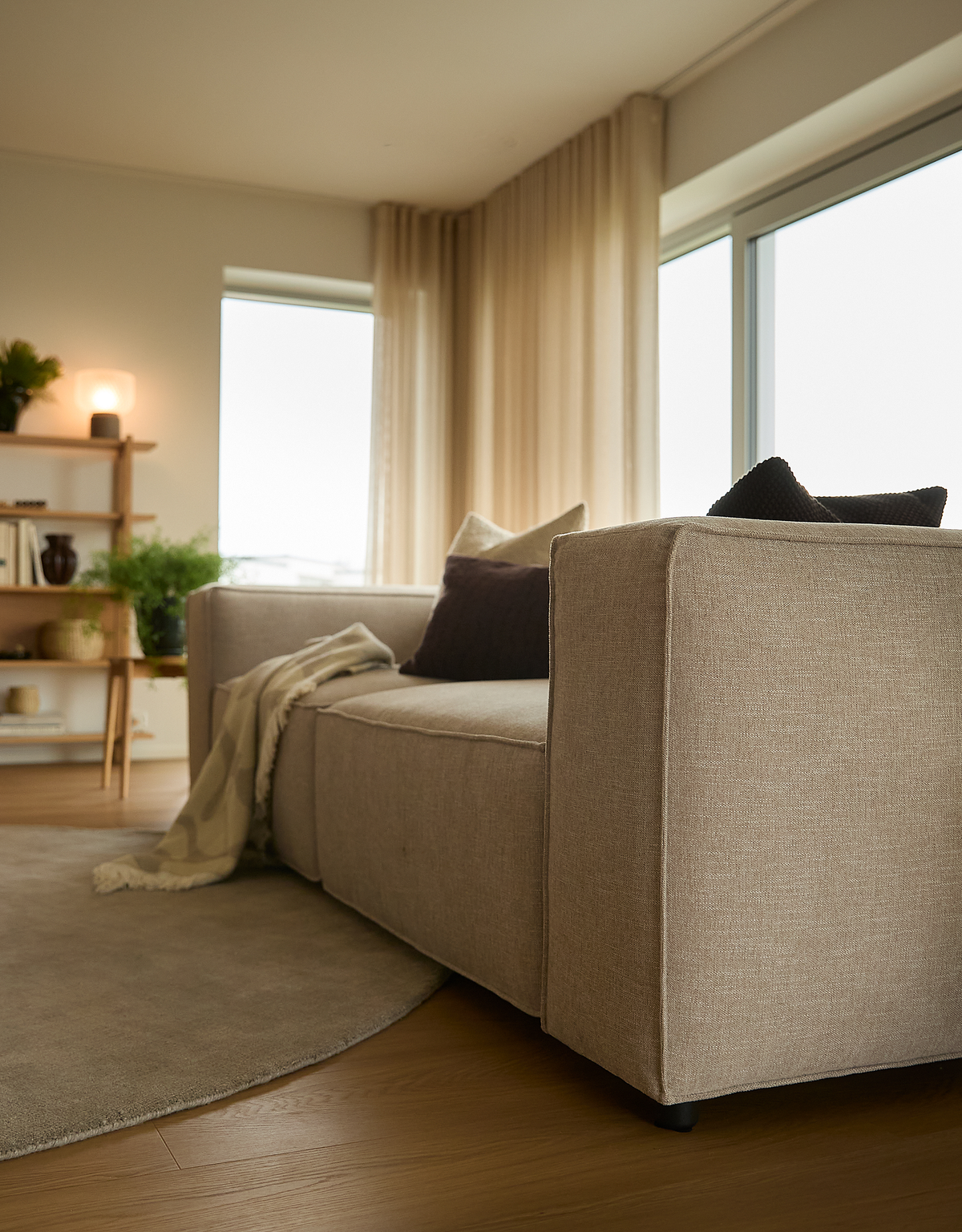 Beige sofa i lyst skandinavisk stue med puder, plaid og store vinduer.