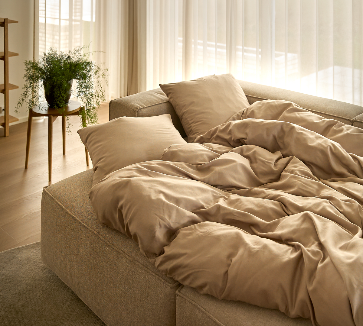 Redt sovesofa med beige, satinlignende sengetøj og puder i blødt dagslys, med plante og lette gardiner i baggrunden.