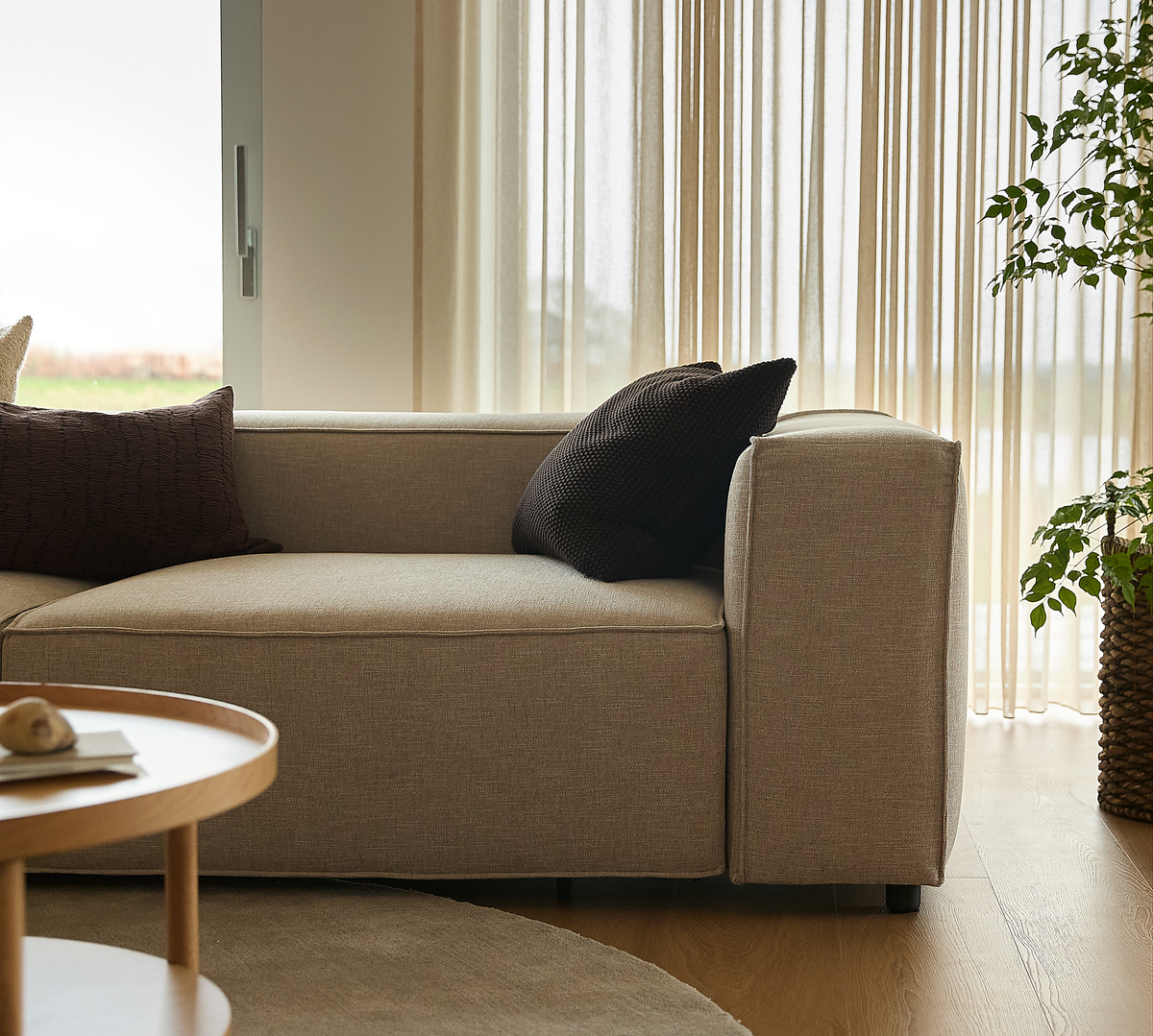 Beige sofa med mørke puder i lys stue med lette gardiner, rundt træbord og grøn potteplante.