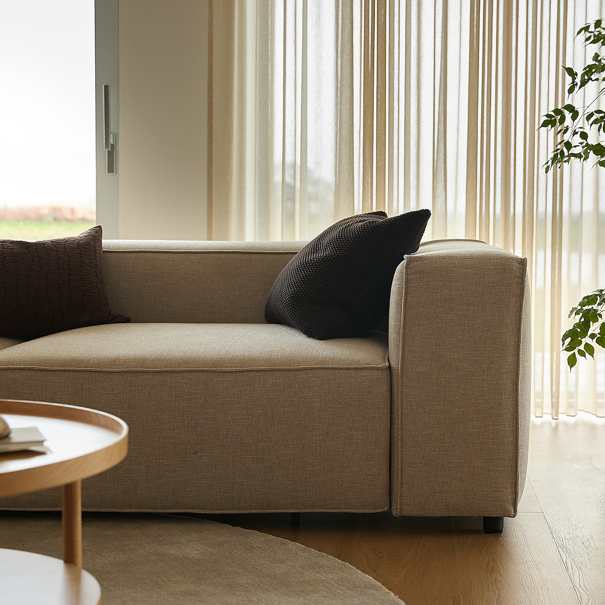 Beige sofa med mørke puder ved lyse, lette gardiner, rundt sofabord i træ og grøn plante i en lys stue.