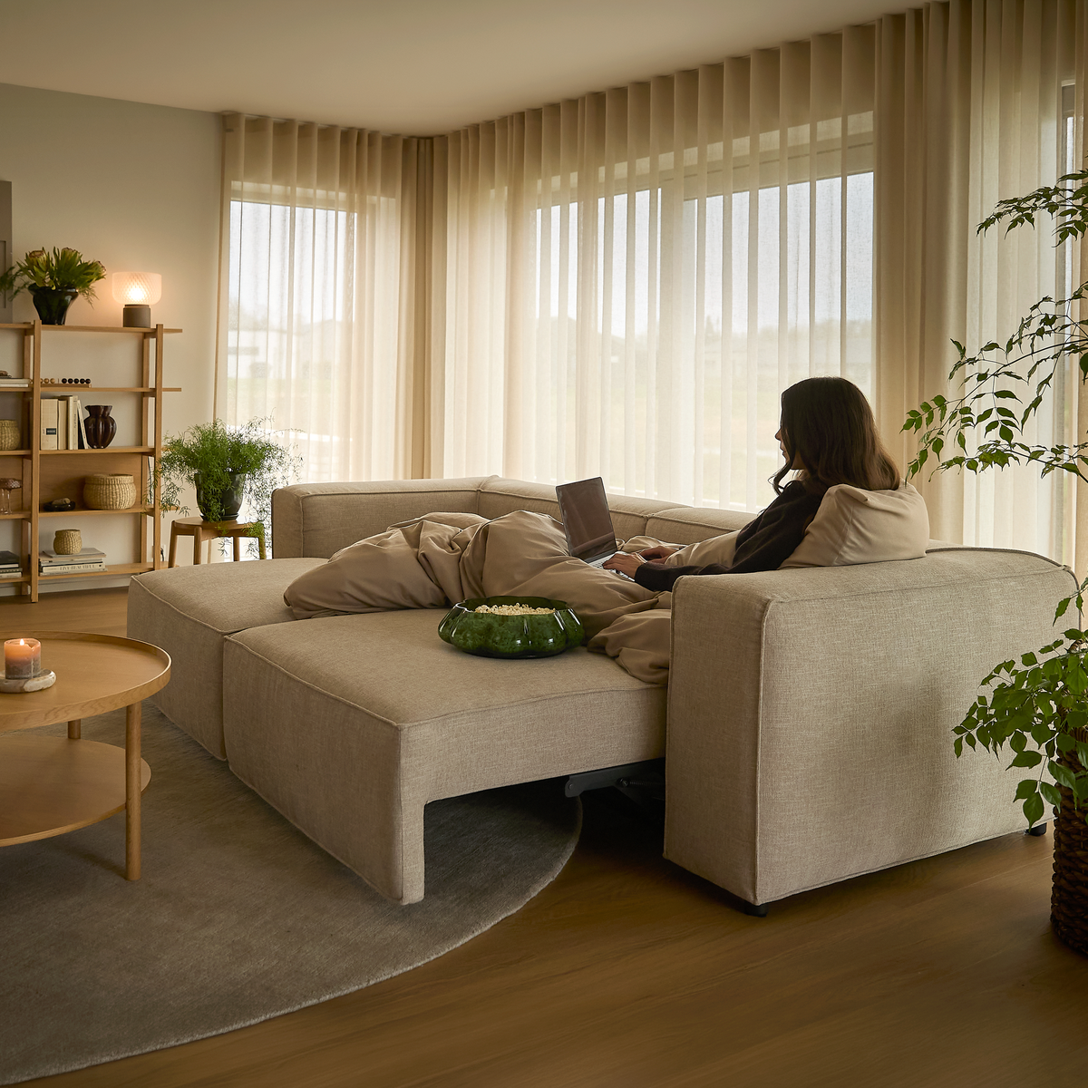Lyst og hyggeligt stuemiljø med beige modulsofa udslået som lounge, person der arbejder på laptop, lette beige gardiner, planter, reol, rundt sofabord med tændt lys og grå tæppe i dagslys.