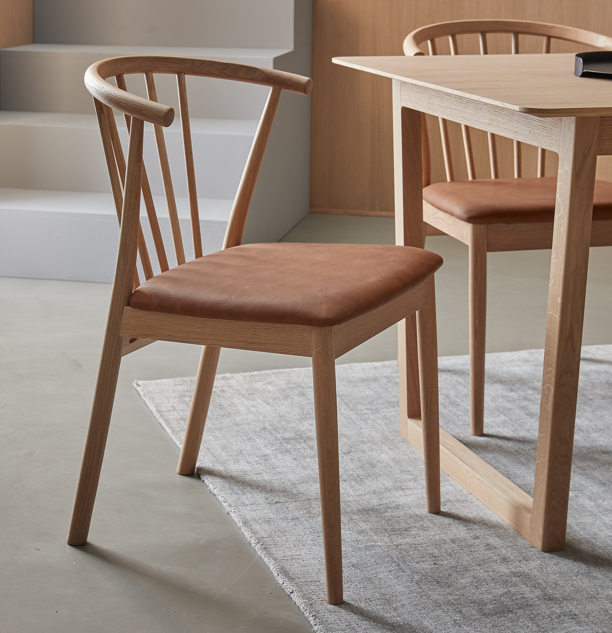 Træstol med brun læderpolstring ved lyst spisebord på gråt tæppe i moderne spisestue.