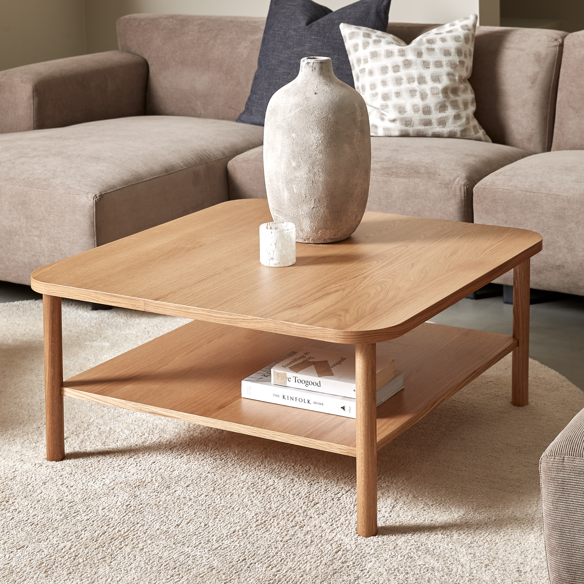 Kvadratisk sofabord i eg med afrundede hjørner, en keramikvase og et lille lys ovenpå samt bøgerne Faye Toogood og The Kinfolk Home på hylden; beige sofa og blød tæppe i en lys stue.