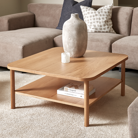 Kvadratisk sofabord i eg med afrundede hjørner, en keramikvase og et lille lys ovenpå samt bøgerne Faye Toogood og The Kinfolk Home på hylden; beige sofa og blød tæppe i en lys stue.