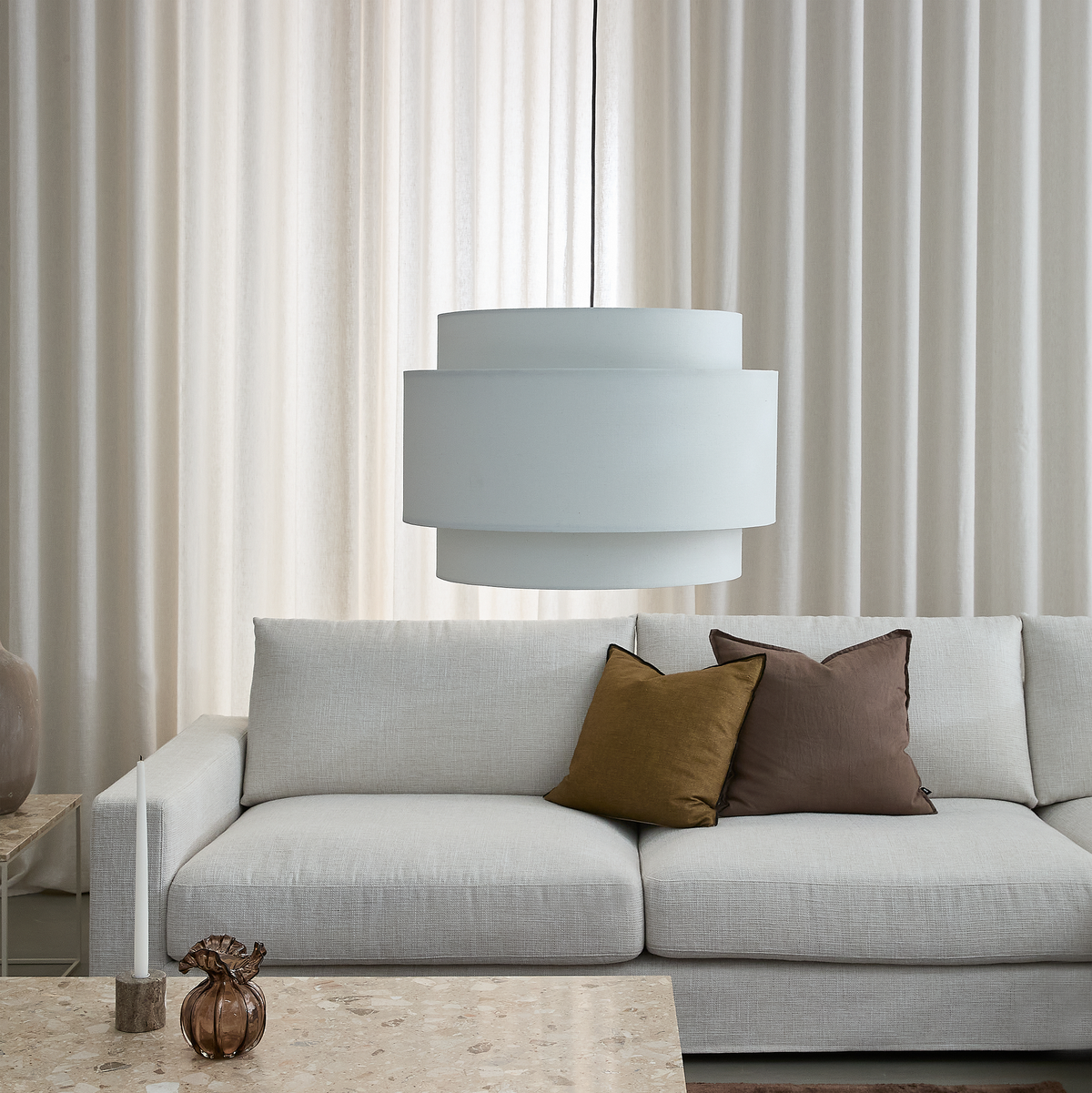 Lys, minimalistisk stue med lysegrå sofa, to puder i brun og oliven, stor hvid pendellampe, lyst stenbord med lys og lille glasvase samt beige gardiner bagved.