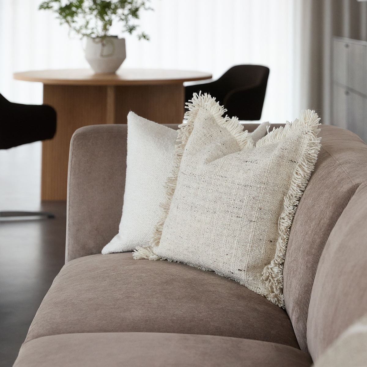 Beige sofa med to lyse, teksturerede pyntepuder, den ene med frynser; i baggrunden et rundt træbord, en sort stol og en hvid vase med grønt foran lyse gardiner.