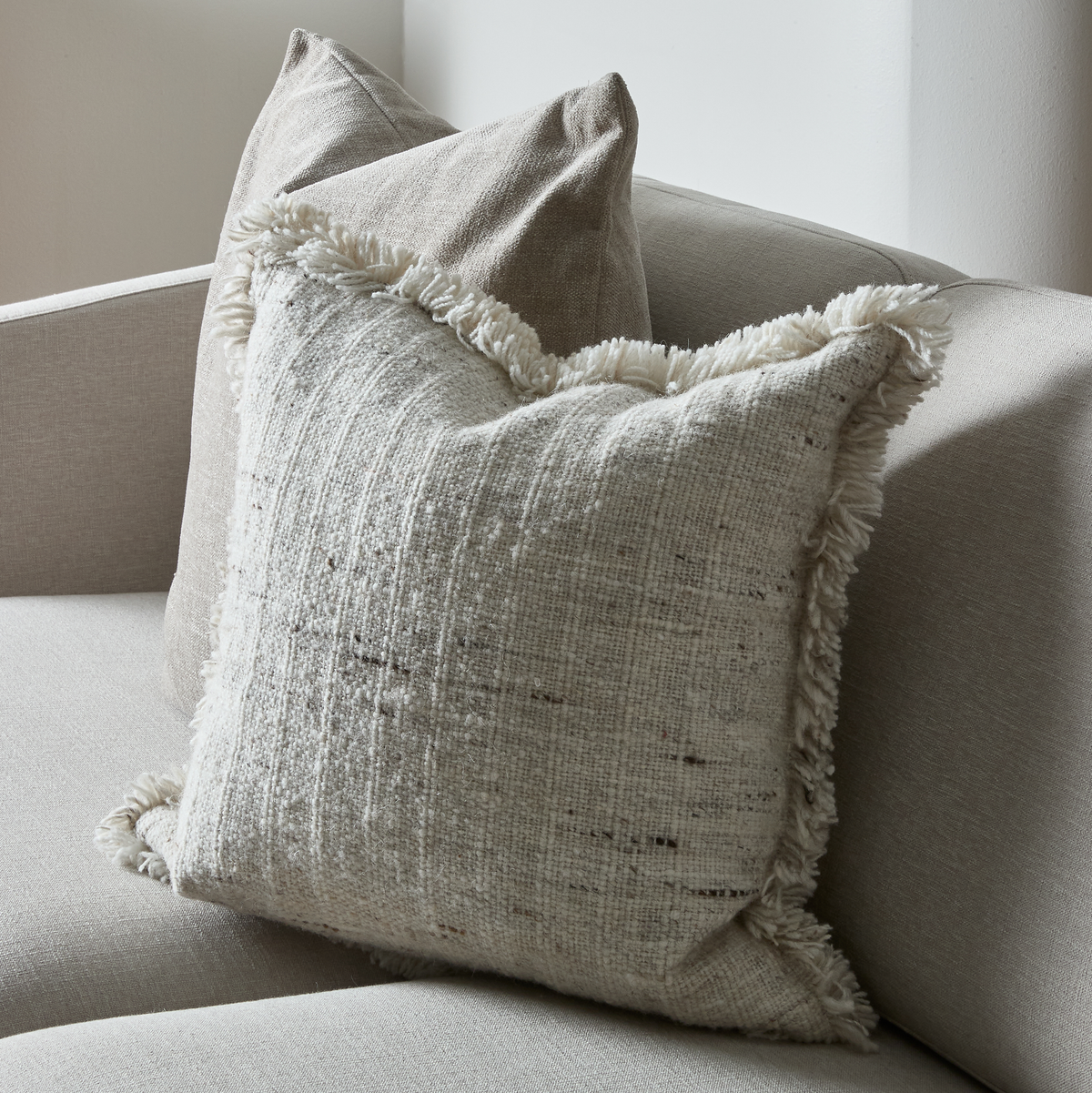 Nærbillede af en struktureret cremefarvet pyntepude med frynser på en beige sofa.