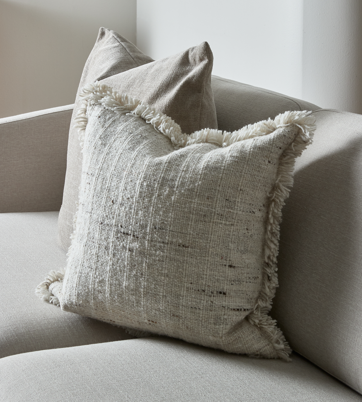 Nærbillede af to neutrale pyntepuder på en beige sofa; forreste pude i offwhite, groft vævet med frynser.
