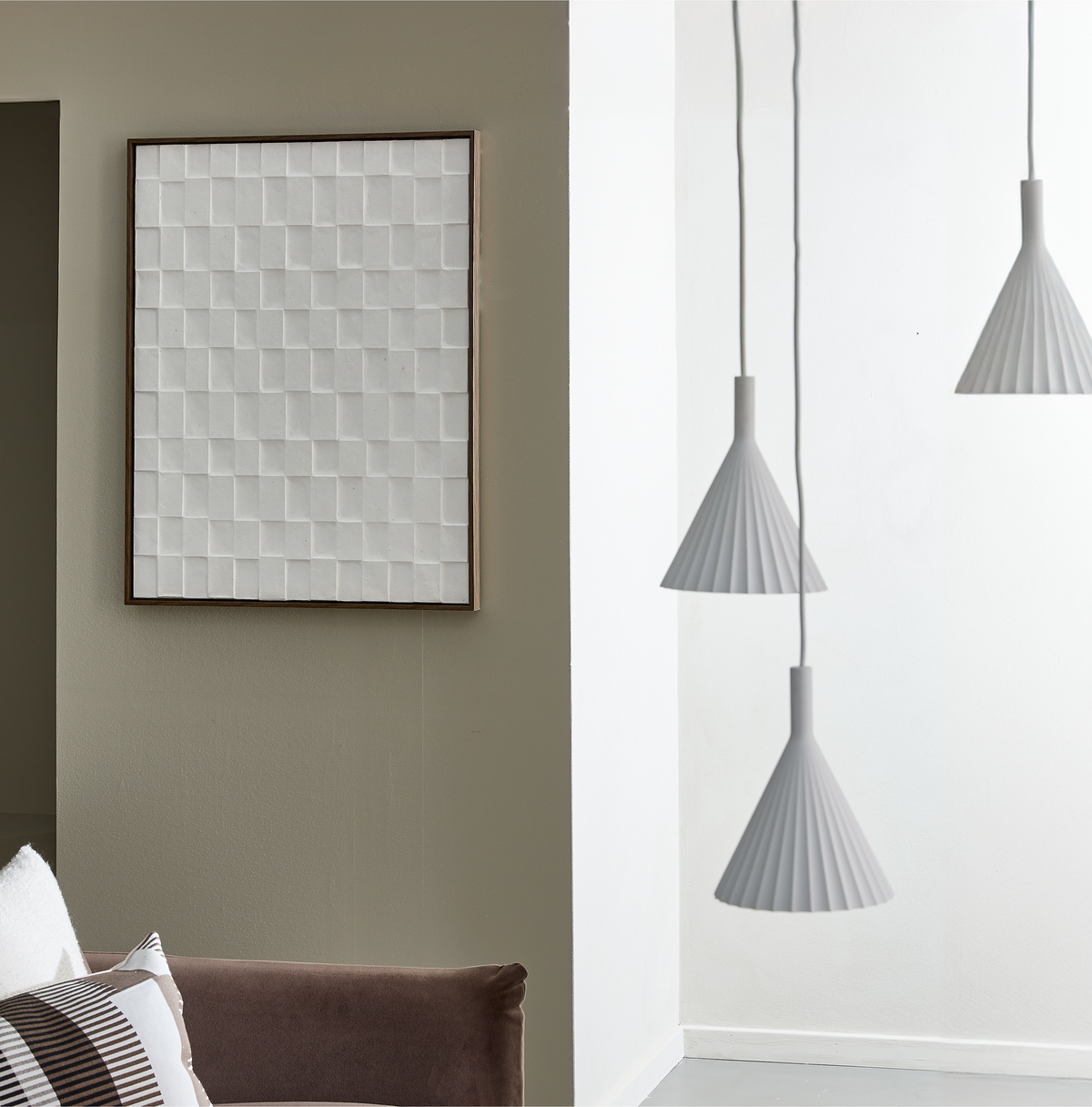 Tre hvide pendellamper foran en lys væg ved siden af en beige væg med indrammet hvid relief, del af sofa med puder.