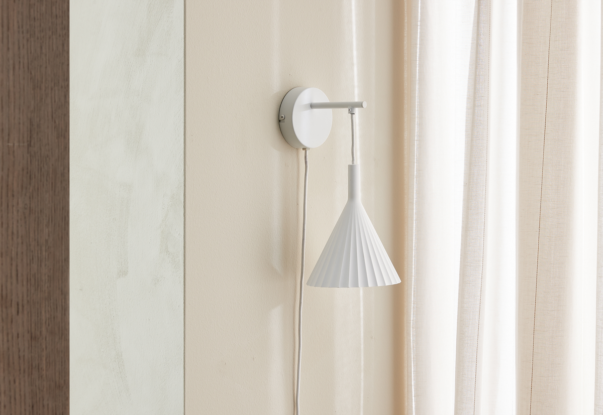 Hvid vægmonteret lampe med plisseret, kegleformet skærm ved siden af lyse gardiner i et neutralt rum.