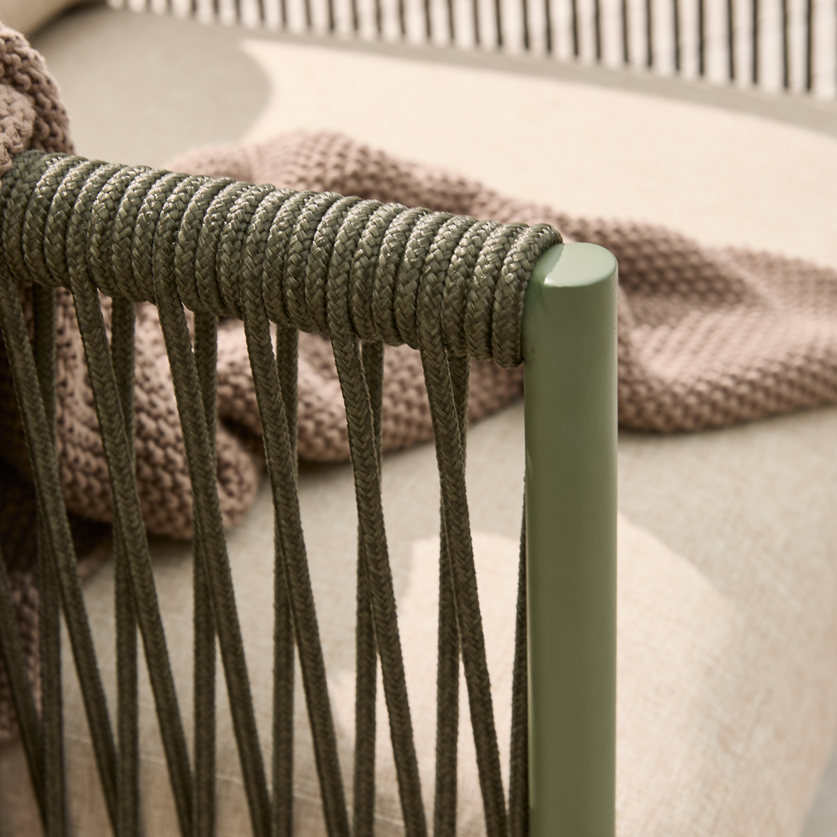 Nærbillede af grøn udestol med flettet rebdetalje, beige hynde og strikket brunbeige plaid.