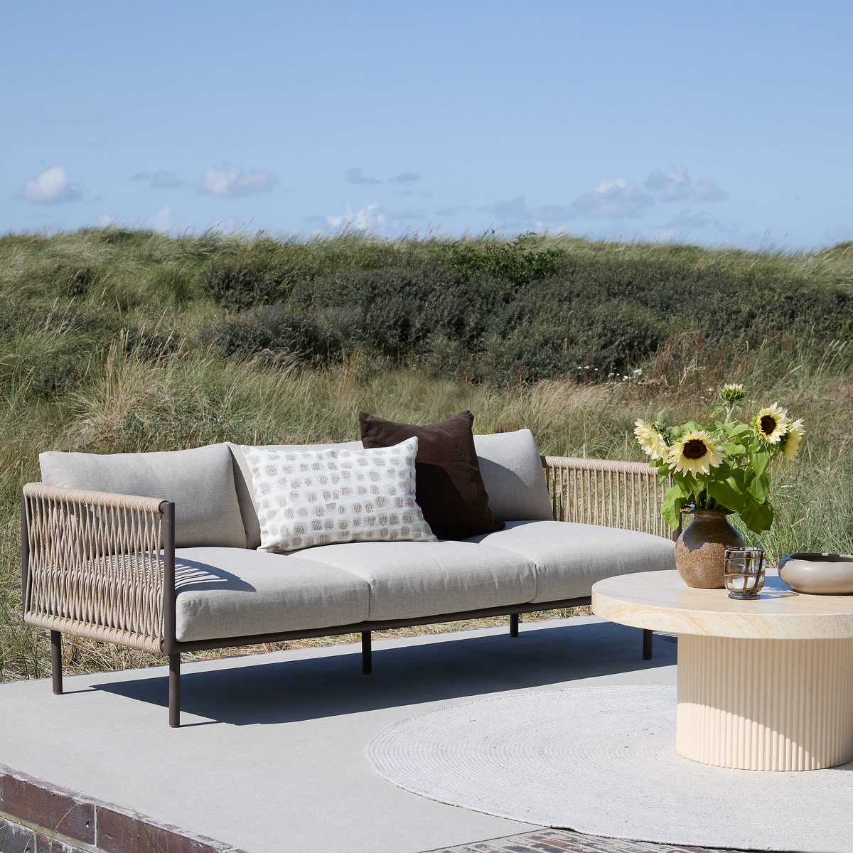 Udendørsscene med beige flettet loungesofa med puder og rundt lyst stenbord med en vase med solsikker på en terrasse, med græsklædte klitter og blå himmel i baggrunden.