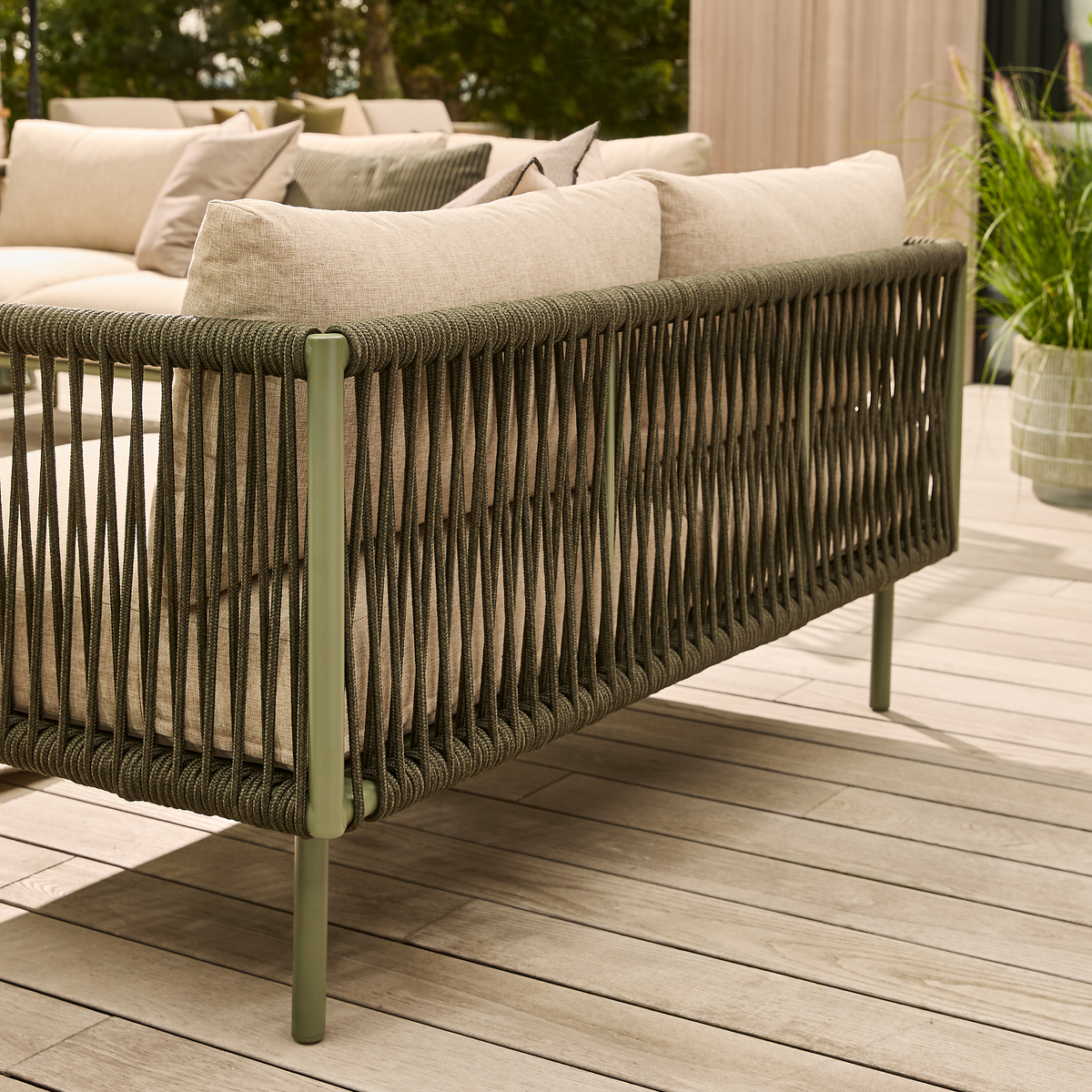 Nærbillede af udendørs sofa med olivengrøn snorre-ramme og beige puder på træterrasse, med potteplante i baggrunden.