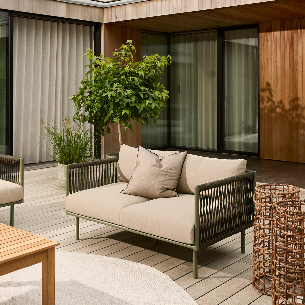 Lys terrasse med beige udendørs sofa med flettsider, kurvelanterner og grønne krukkeplanter ved træfacade og glas-skydedøre.