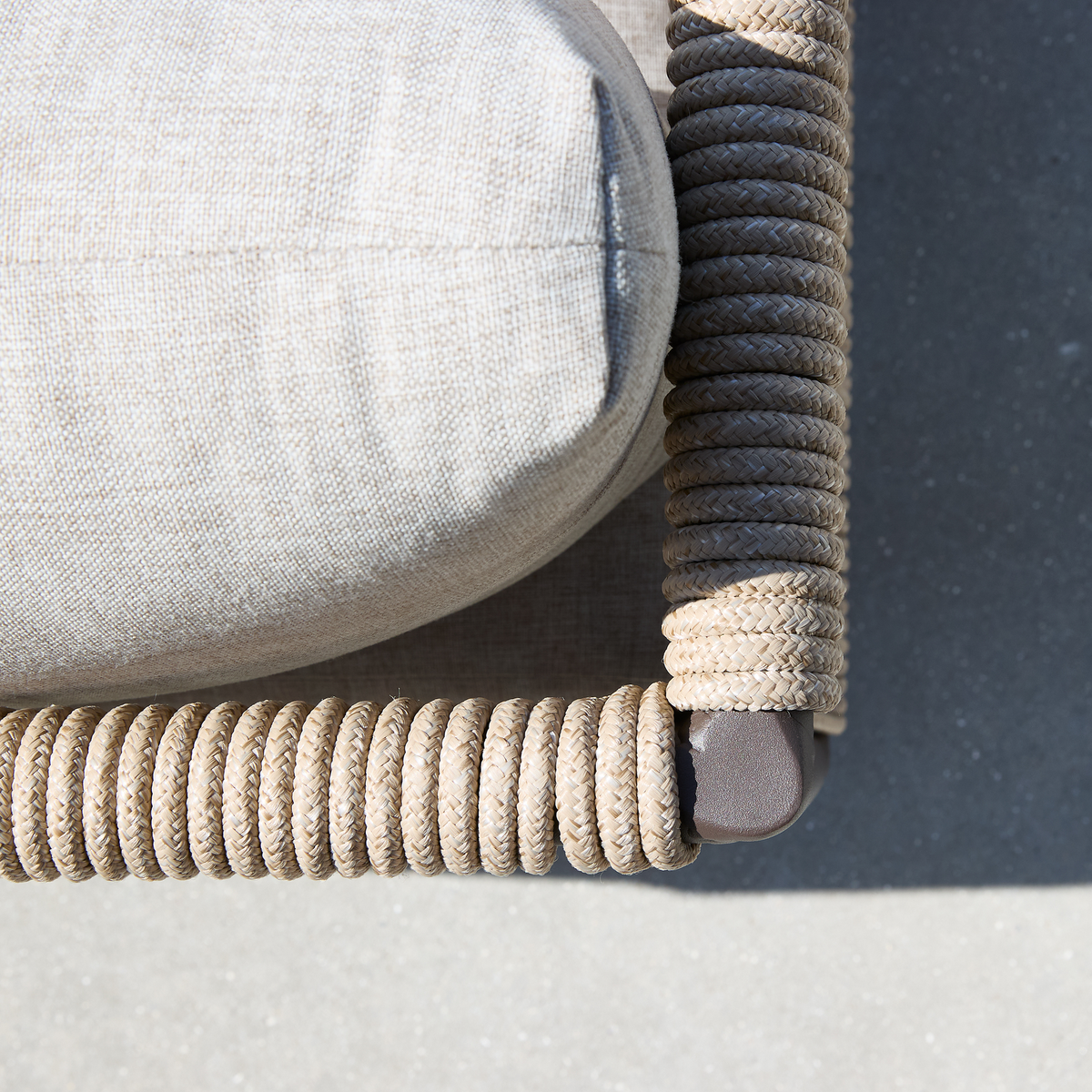 Nærbillede af hjørnet på en udendørsstol med lys beige pude og tykt flettet reb omkring rammen på grå beton.