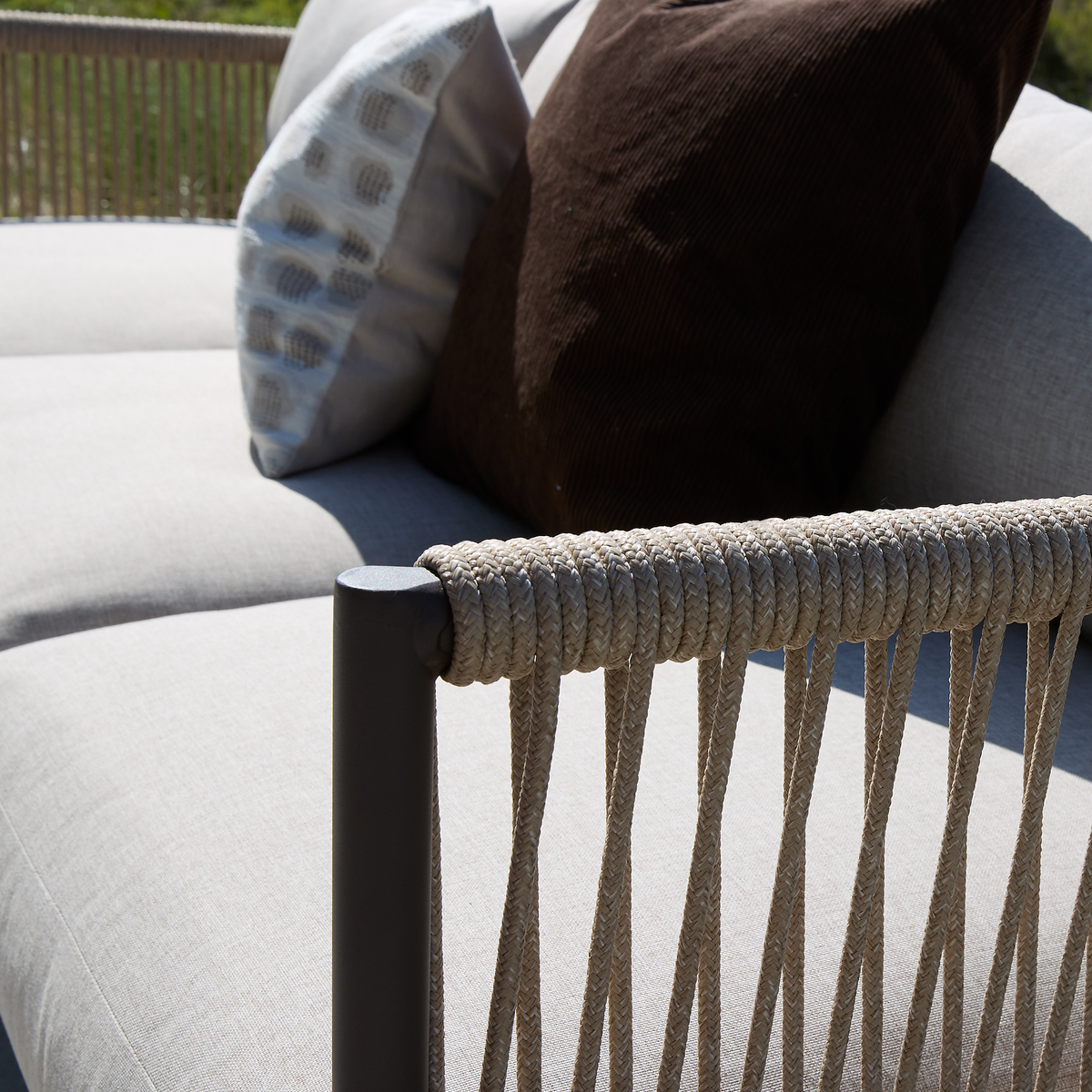 Nærbillede af udendørs sofa med beige hynder, armlæn viklet i reb og to pyntepuder – en brun og en lys mønstret – i sollys.