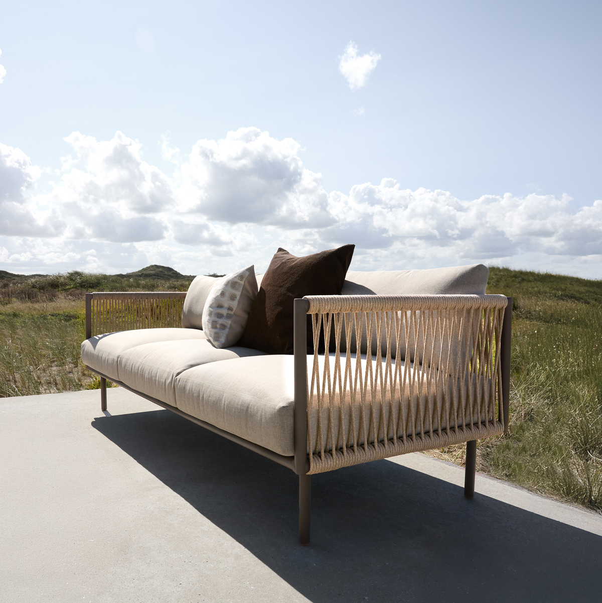 Udendørs sofa med flettede sider og beige hynder, pyntet med brune og mønstrede puder på en terrasse med klitter og blå himmel med skyer.