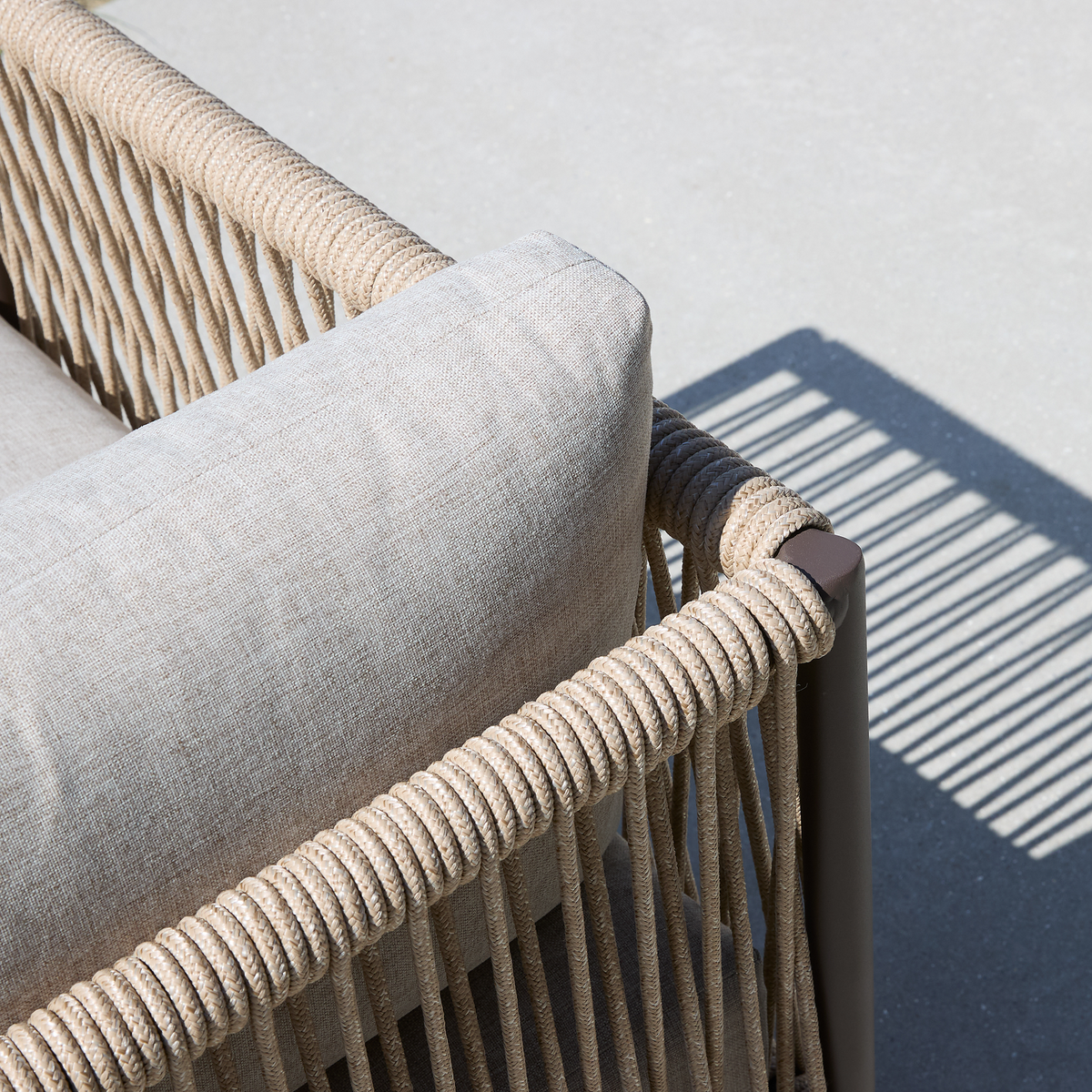 Nærbillede af hjørnet på en beige udendørs sofa med rebflettet ramme og lys pude, der kaster stribet skygge på beton.