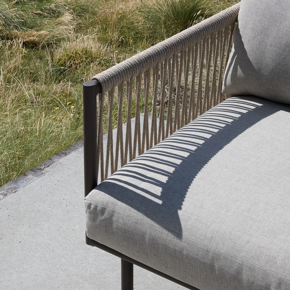 Nærbillede af udendørs sofa med reb i ryglænet, der kaster stribet skygge på lysgrå hynde på betonterrasse ved højt græs.