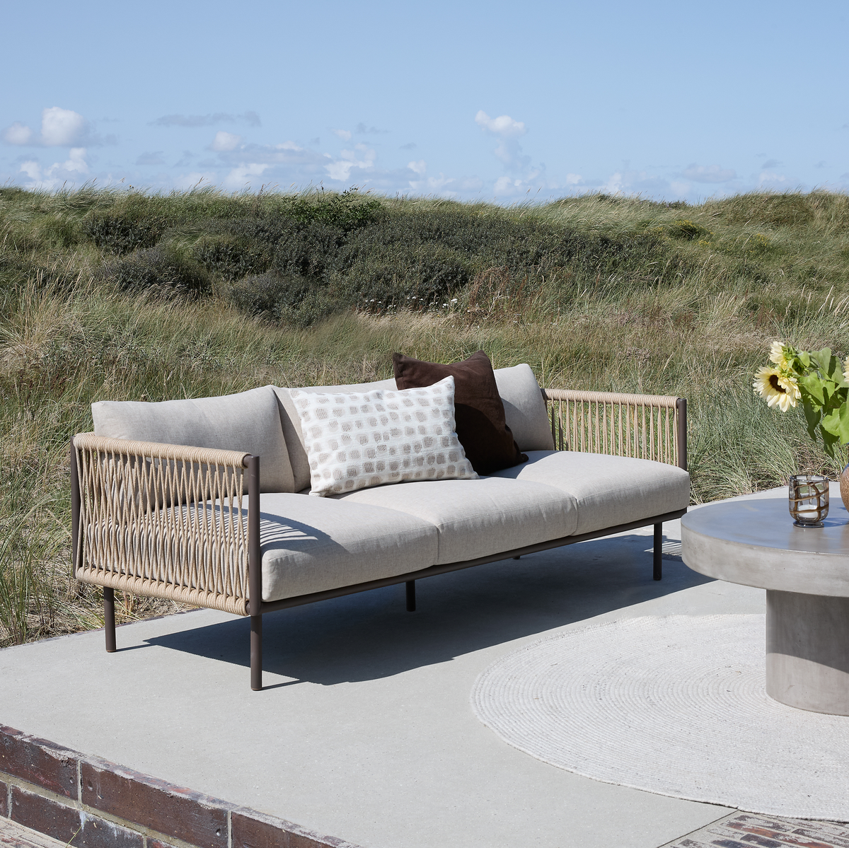Udendørs beige sofa med puder på en betonterrasse ved siden af rundt betonbord med vase og solsikke, med græsklædte klitter og blå himmel i baggrunden.