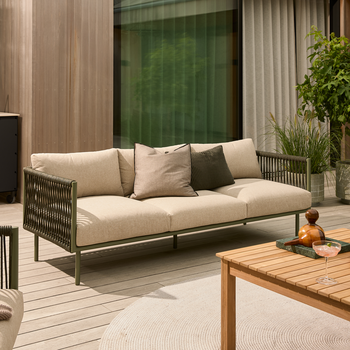 Beige udendørs sofa med flettede armlæn og mix af puder på en moderne terrasse ved siden af et lameltræsbord, planter og et rundt tæppe i dagslys.