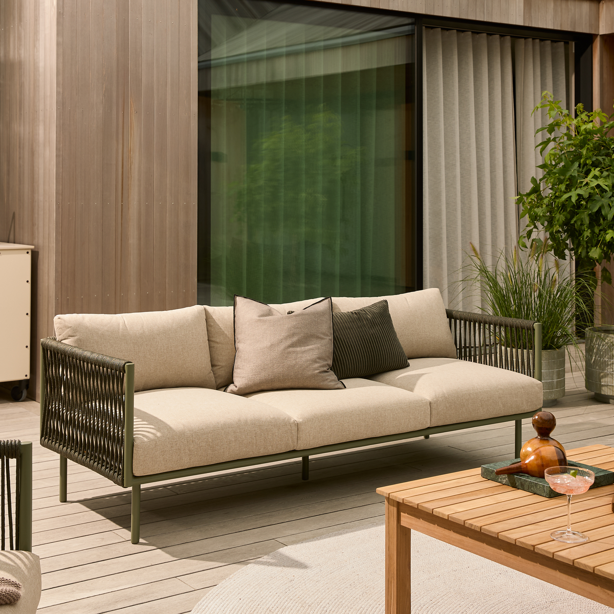 Moderne udendørs beige sofa med puder på en træterrasse, ved siden af et træbord og grønne planter i dagslys.