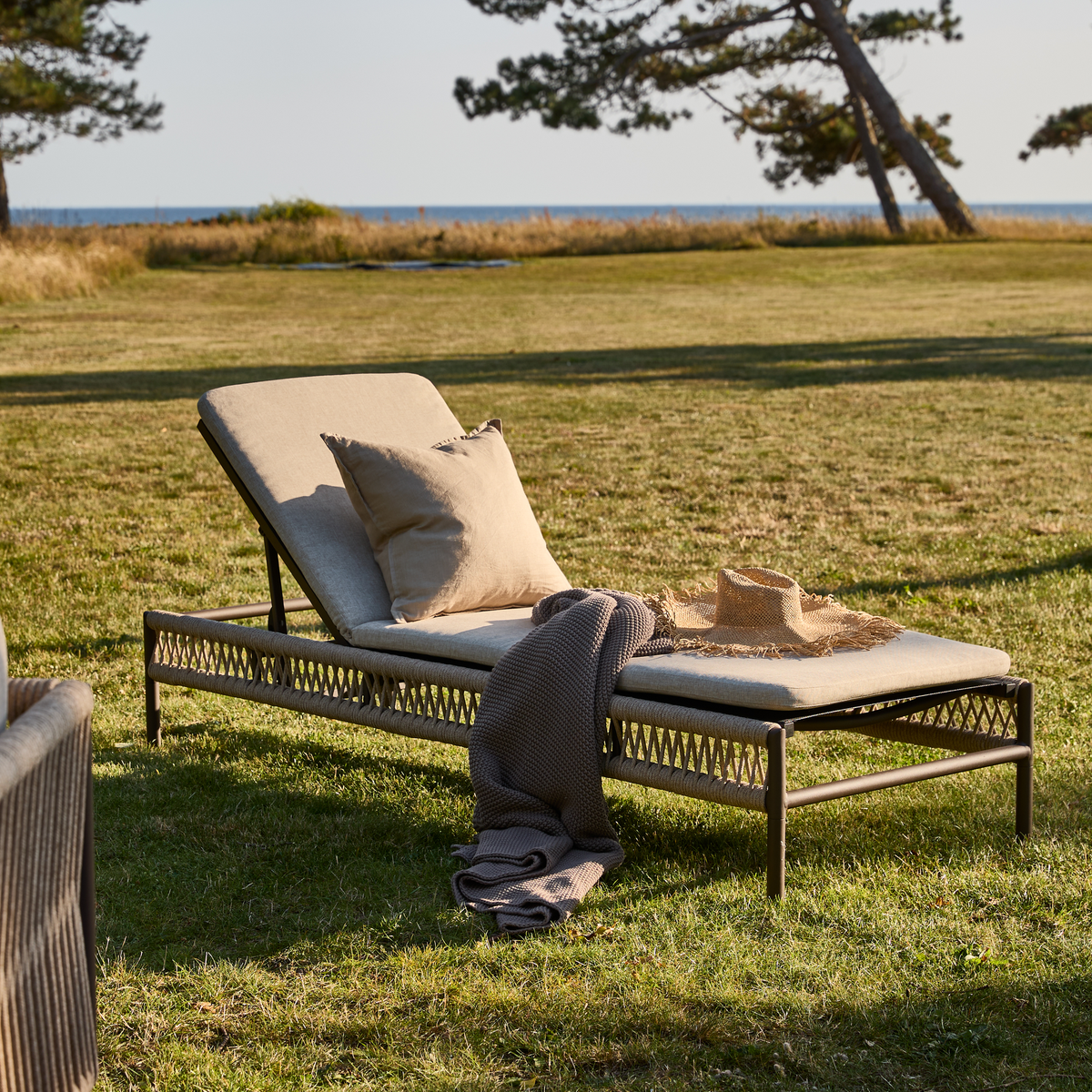 Solseng på græsplæne ved havet med beige madras, pude og plaid samt stråhat.