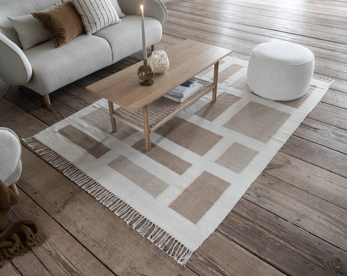 Lyst stueinteriør med beige‑hvidt geometrisk tæppe, sofabord i træ med tændt lys og bøger, sofa og rund puf på trægulv.