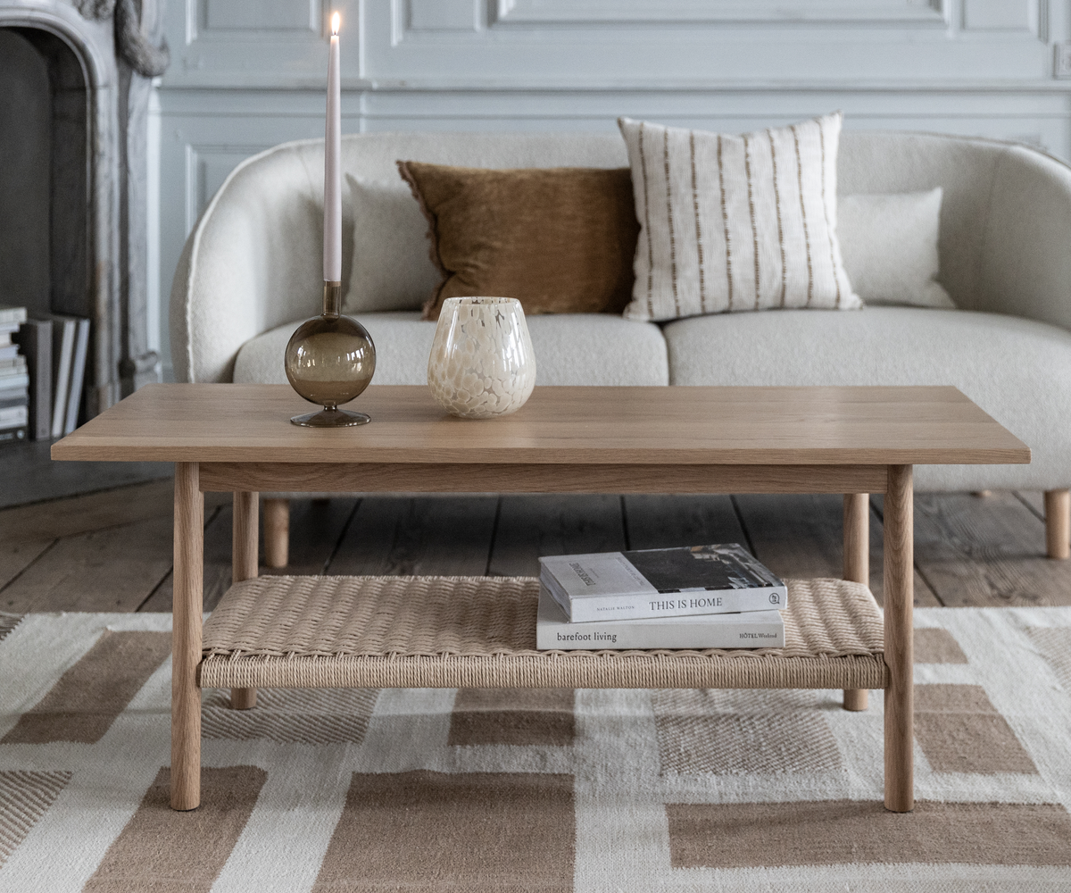 Lyst stue med sofabord i eg; på bordet står et tændt stearinlys i brun stage og en lille vase; på hylden ligger bøgerne ”THIS IS HOME” og ”barefoot living”; beige sofa med puder og neutral mønstret tæppe.