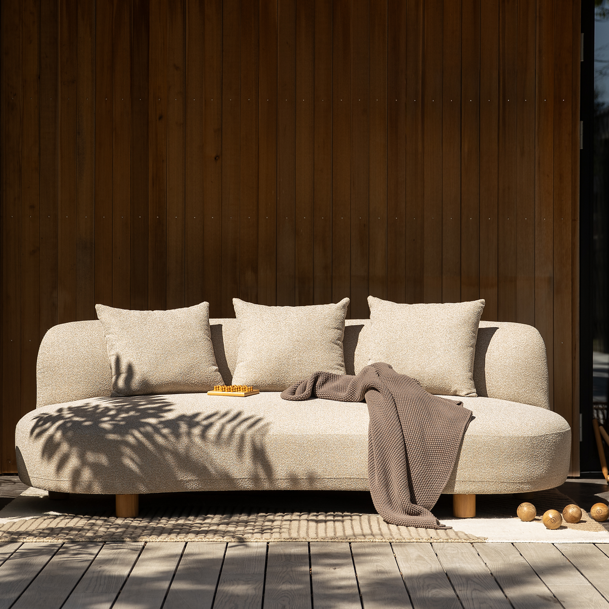 Beige udendørs sofa med tre puder, et brunt plaid og et lille skakbræt på en træterrasse foran en væg med træpaneler.