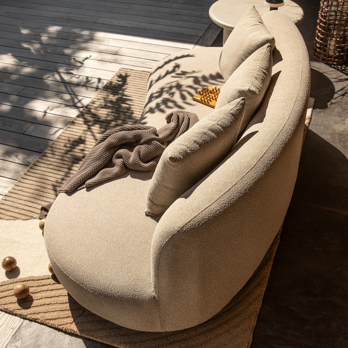 Beige, buet sofa på solrig træterrasse med stribet neutral tæppe, puder og brun strikket plaid; dekorative trækugler og plettet skygge fra planter.