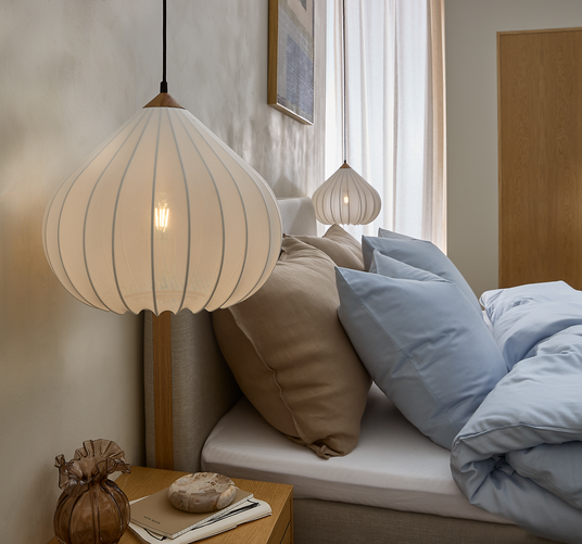 Soveværelse med hvide hængelamper over en seng med beige og lyseblå puder; træ-natbord med glasvase og bog i blødt dagslys gennem lyse gardiner.
