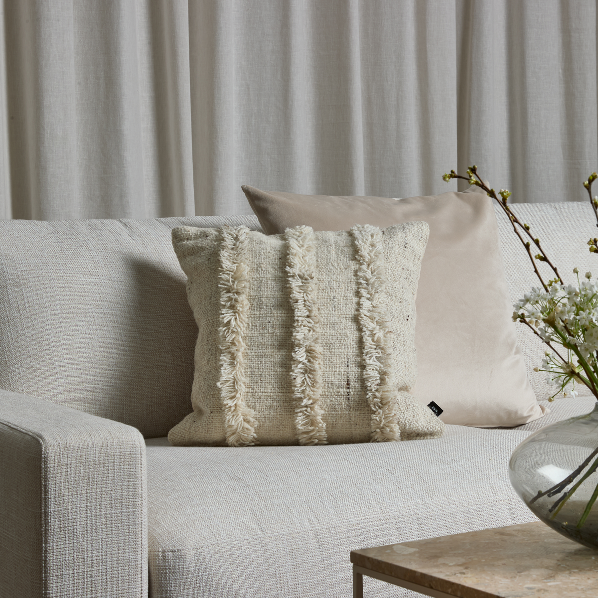 Beigefarvet sofa med to pyntepuder, den ene med frynsede striber; lyse gardiner bagved og glasvase med hvide blomster på et stenbord.