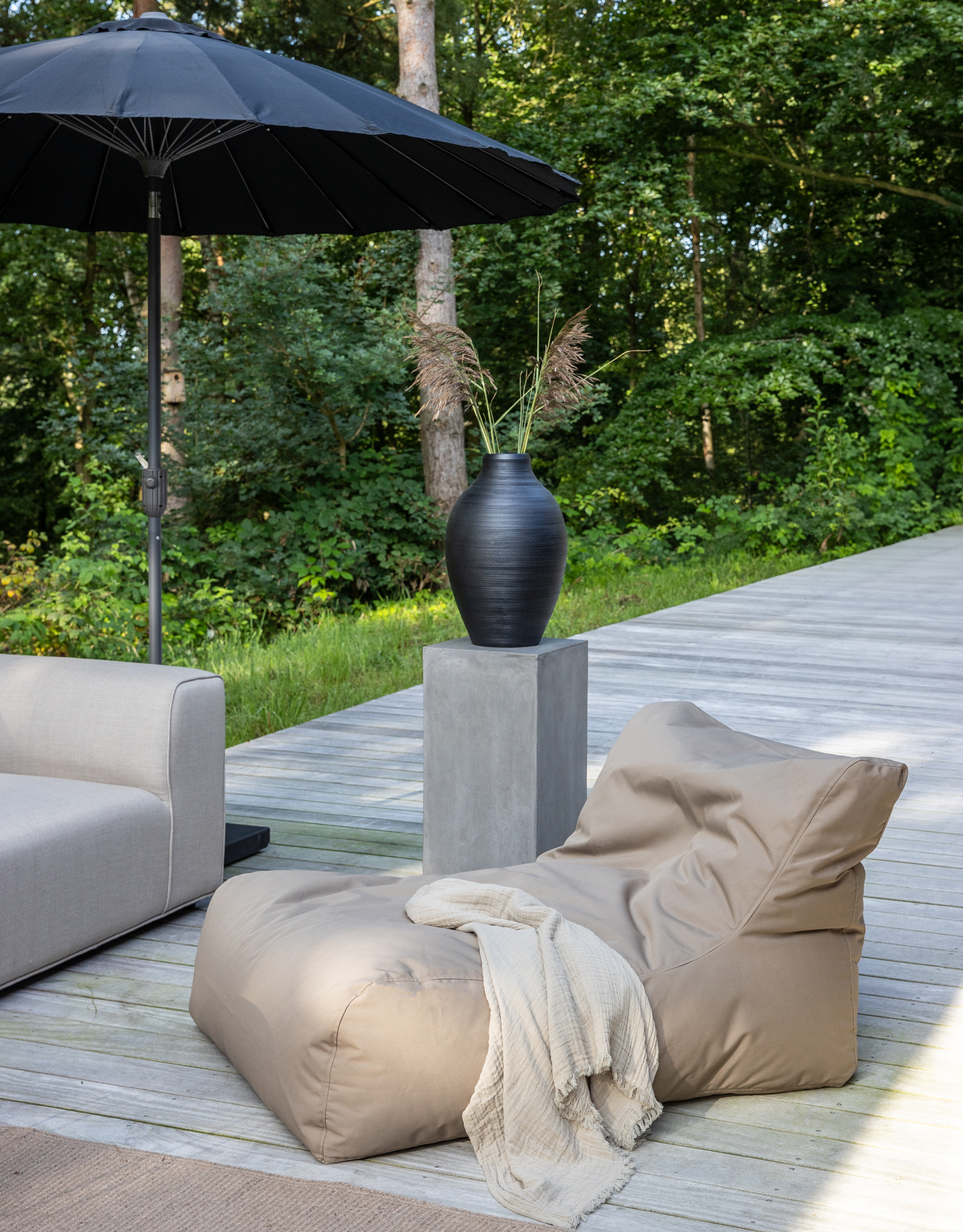 Udendørs loungemiljø på trædæk med beige sækkestol, lys plaid, sort vase på betonsokkel og stor sort parasol ved grøn skov.