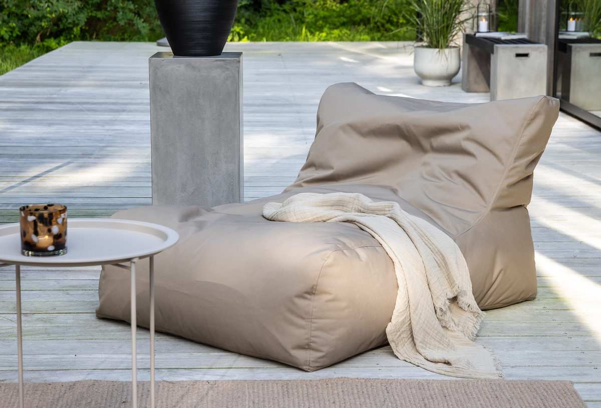 Beige lounge-sækkestol på træterrasse med lys plaid, rundt sidebord med lysestage og krukker med grønne planter i dagslys.