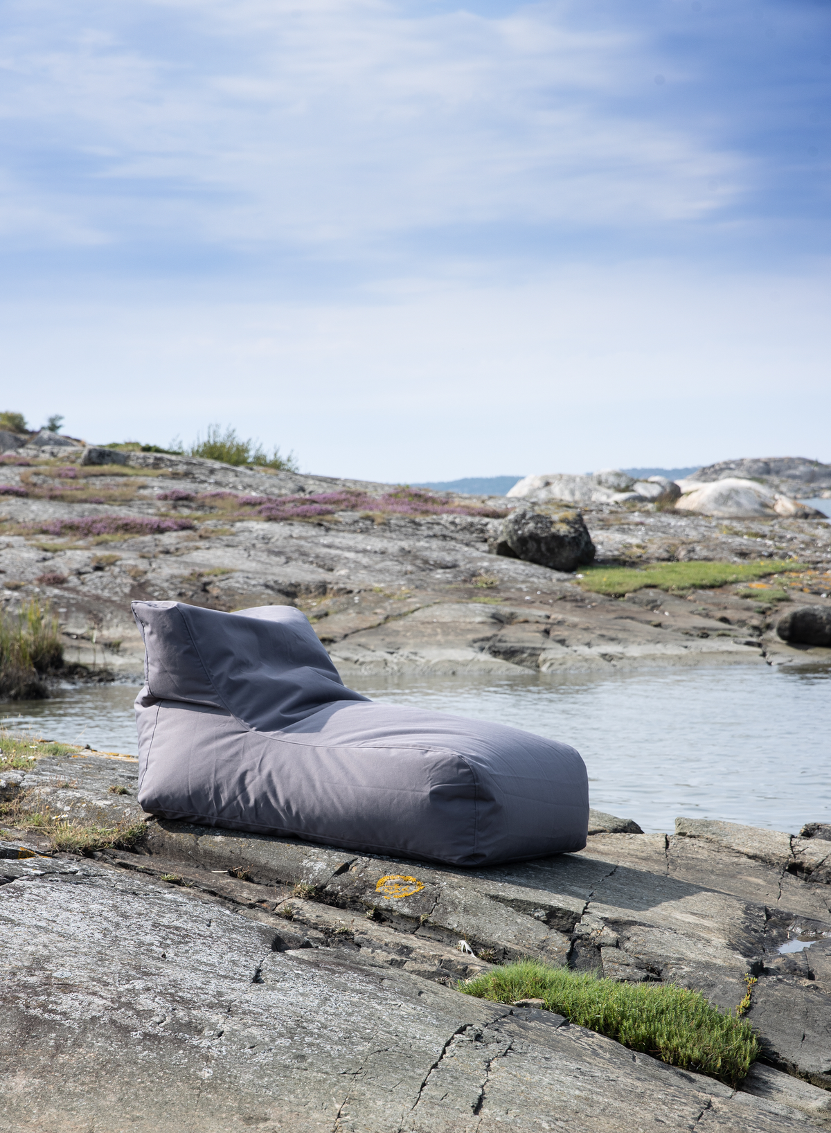 Grå loungestol i sækkestolsform på klipper ved vandet under blå himmel.